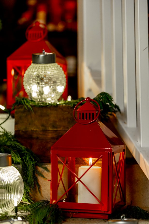 This red lantern has been filled with a candle and lit up to line a stairway leading to a front porch. Greenery is strewn along the steps surrounding the lantern and intertwining around homemade Christmas ornaments. The ornaments are made from vintage light globes.