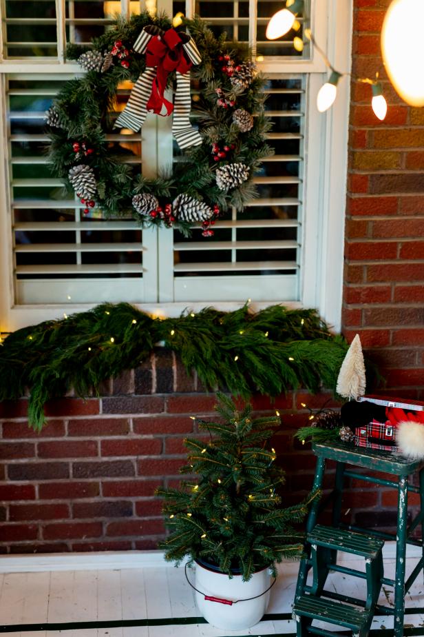 The front porch of this home has been decorated using a mini potted Christmas tree, a wreath with pine cones and ribbon and boughs of greenery. The decorations are lit up using mini string lights and the glowing ambience added is beautiful against the brick home.