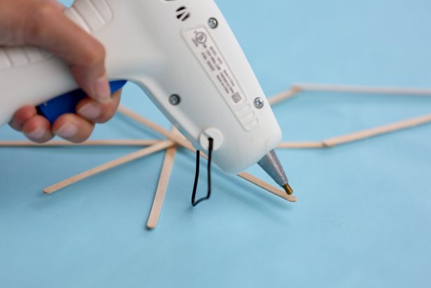Hot glue is used to attach coffee stirrers to one another to make a star Christmas decoration. The minimalistic decor piece will be displayed in a window to cast a slender silhouette.