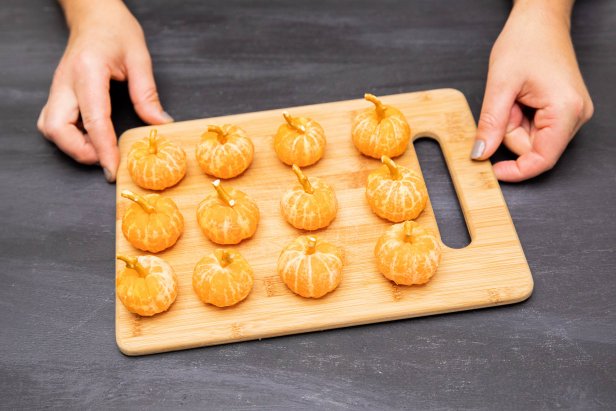These mini pumpkins are crafted using clementine oranges that have been peeled and had broken pretzel pieces stuck in the center to look like stems. The oranges are served up on a wooden cutting board.