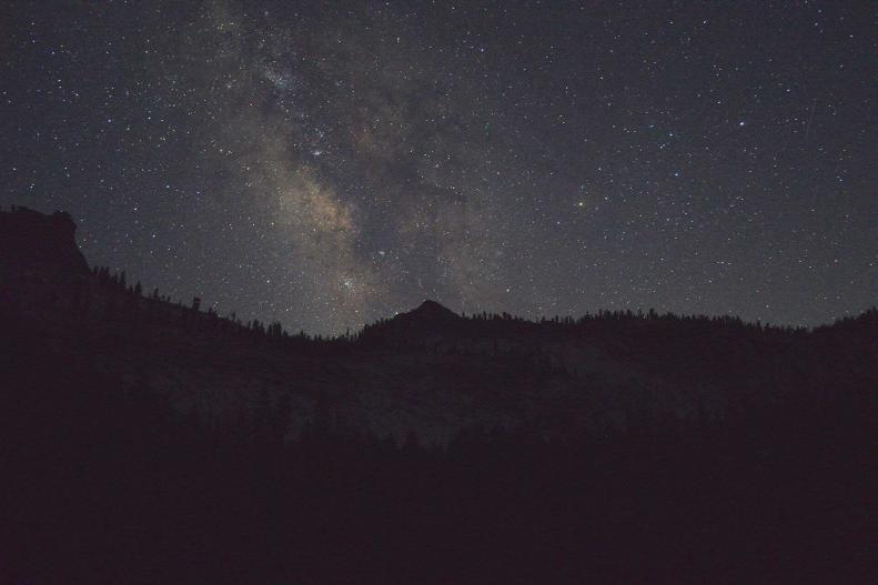 Stars and cosmic dust over darkened mountains and trees.