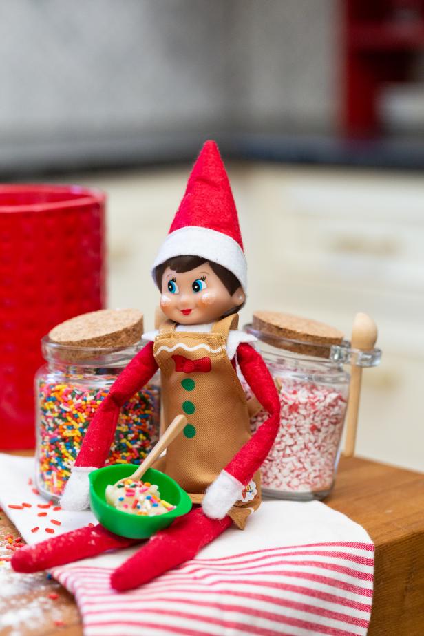 A toy elf holds a tiny green bowl with flour and sprinkles. 