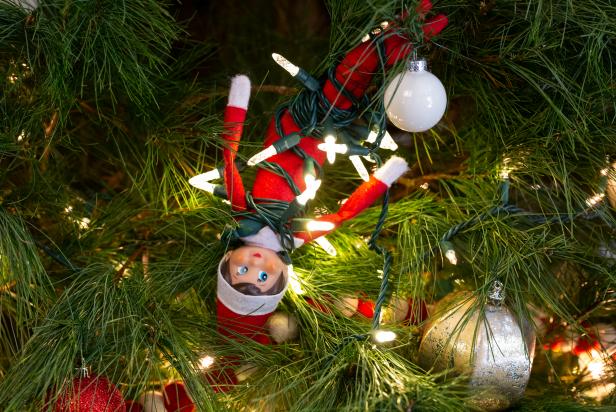 A toy elf wrapped in Christmas lights hangs upside down in a tree.