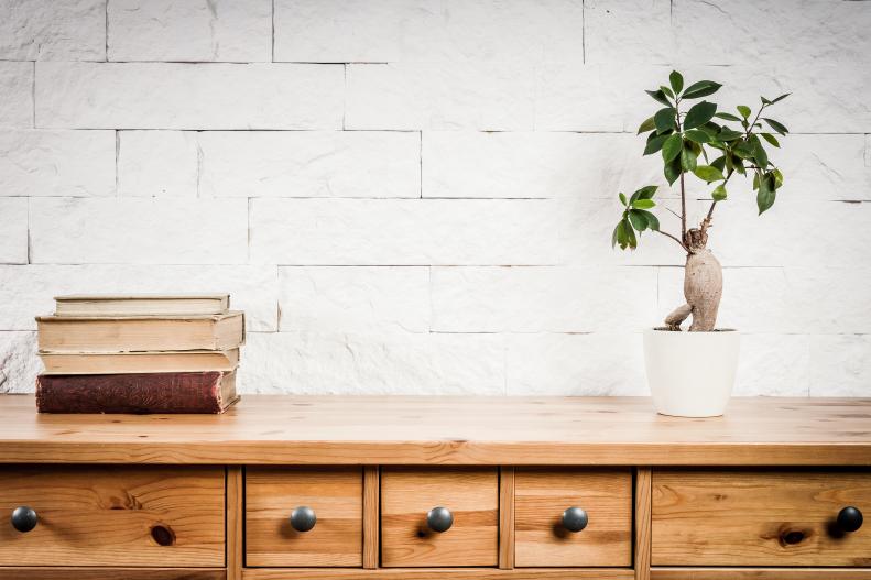 An unfinished wood chest of drawers sits against a painted white brick wall.