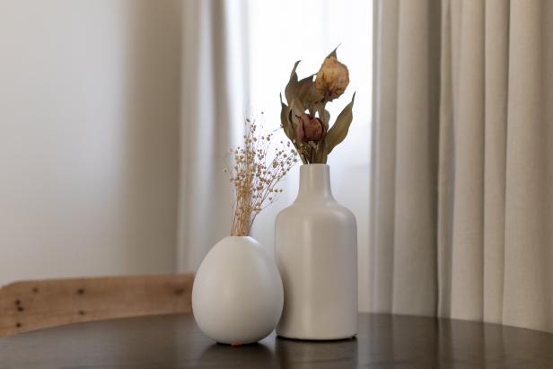 Two modern white vases on a table feature dried flowers.