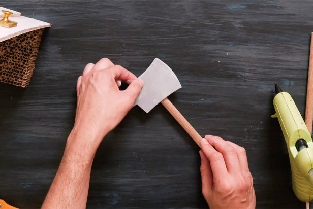 Using a crafting knife, cut a small piece of gray crafting foam into two axe blade shapes so it’s double-sided. Now, cut a wooden dowel into two even pieces. Place one piece of the wooden dowel in-between the two axe blade shapes and use a low temp hot glue gun to adhere together. Next, wrap the remaining dowel tightly with the large furry pipe cleaner. Be sure to use a furry pipe cleaner that closely matches your cat’s fur.