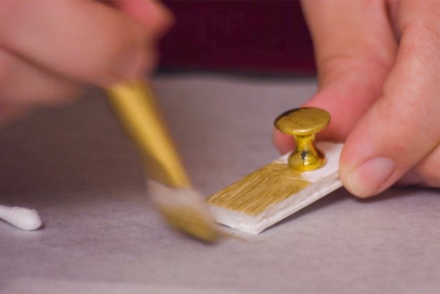 With a crafting knife, cut a small rectangle from the remaining foam board. Next, grab your tack and add a bit of glue using a low temp hot glue gun. Stick the tack into the foam board piece. Last, paint the entire piece with gold acrylic paint and glue the piece to the door.