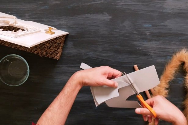 Using a crafting knife, cut a small piece of gray crafting foam into two axe blade shapes so it’s double-sided. Now, cut a wooden dowel into two even pieces. Place one piece of the wooden dowel in-between the two axe blade shapes and use a low temp hot glue gun to adhere together. Next, wrap the remaining dowel tightly with the large furry pipe cleaner. Be sure to use a furry pipe cleaner that closely matches your cat’s fur.