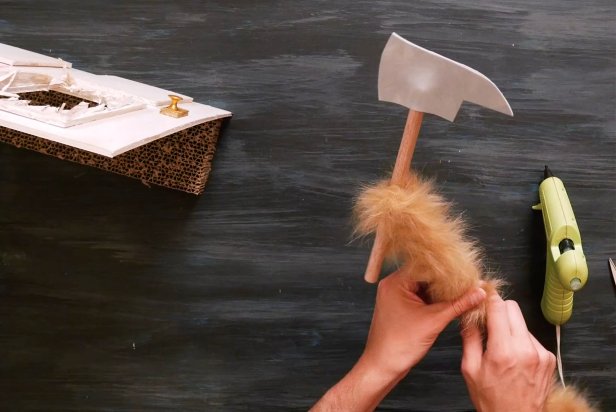 Using a crafting knife, cut a small piece of gray crafting foam into two axe blade shapes so it’s double-sided. Now, cut a wooden dowel into two even pieces. Place one piece of the wooden dowel in-between the two axe blade shapes and use a low temp hot glue gun to adhere together. Next, wrap the remaining dowel tightly with the large furry pipe cleaner. Be sure to use a furry pipe cleaner that closely matches your cat’s fur.