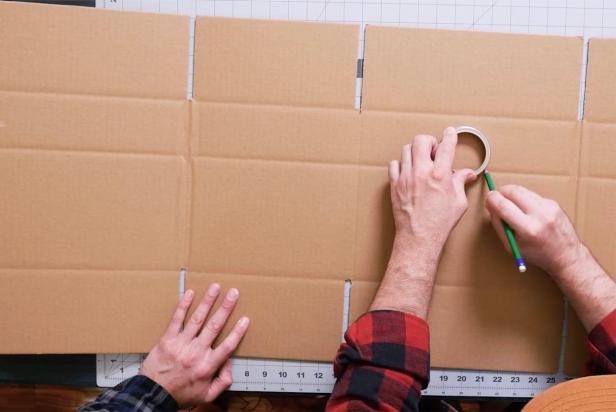 Open up a cardboard box and use a craft knife to slice the seal open so that it lays flat on the work area. Next, using a mason jar lid as a guide, trace a circle with a pencil on the upper part of one side of the box to make the viewing circle. Then, on the back side of the box, trace a larger skull shape to create the larger viewing area. Use a craft knife to cut out the marked pieces.
