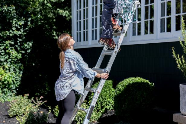 This person is seen holding a ladder to ensure safety as another person installs Christmas lights on the exterior of a home.
