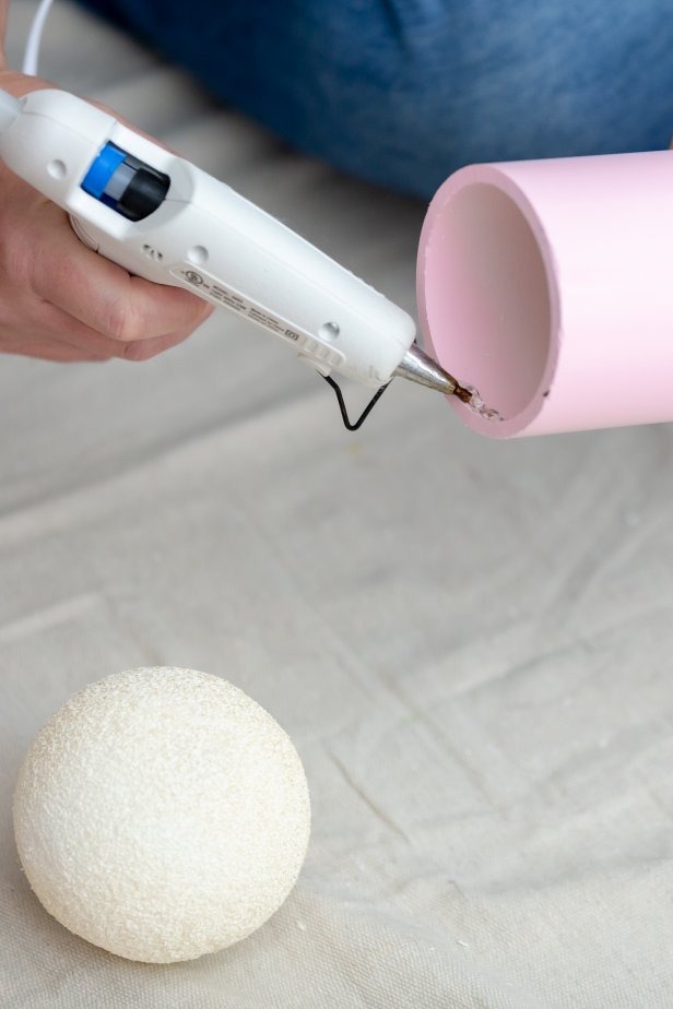 Hot glue is used to attach a small foam ball to a PVC pipe to resemble a hand on an arm of a giant DIY nutcracker Christmas decoration.