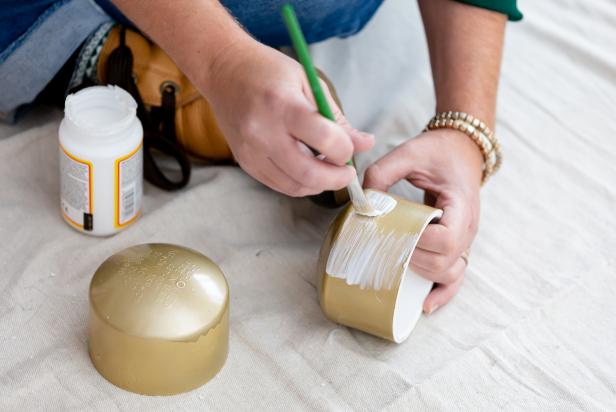 Decoupage glue is brushed onto a PVC pipe cap that has been painted gold in order to apply a layer of gold glitter overtop the paint.