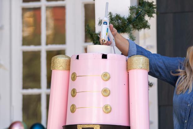 Hot glue is used to secure a large foam ball head to the top of a giant, life-sized nutcracker Christmas decoration made from PVC and plastic planters.