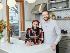 Two Men Stand Smiling for Camera in Kitchen Leaning Against Island 