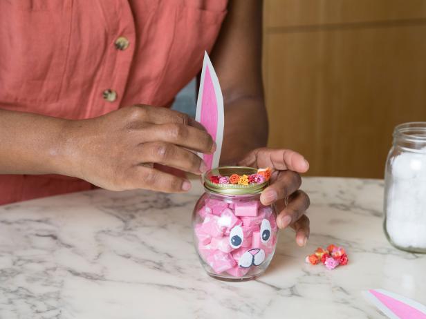 Ears cut out of printer paper are glued to the back of a glass jar decorated to look like a bunny and filled with candy.