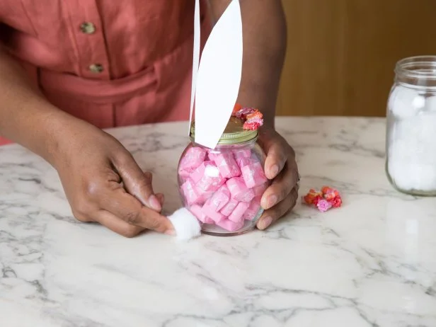 A cotton ball is being glued to the back of a glass jar decorated to look like a bunny and filled with pink candy.