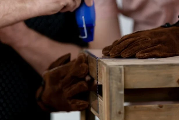 Prepare for the heat gun by putting on heat resistant gloves. Apply heat slowly by waving the heat gun back-and-forth along the edge evenly. As the plastic starts to heat and bend, lightly push down and manipulate the bend to a 90 degree angle using the crate as a guide. Let the acrylic sheet cool before starting the next bend.