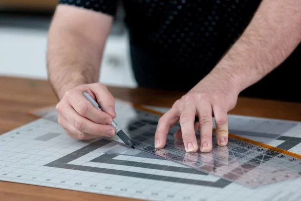 Prepare one of the 8’’x10’’ acrylic sheets by placing it on a cutting mat. Tip: Be sure to leave the plastic covering on the sheet to protect it from any scratches. Then, find the center of the sheet using a metal ruler. Measure 2’’ down each side of the sheet to create a triangle for the bird house peak and mark the triangle with a pen.