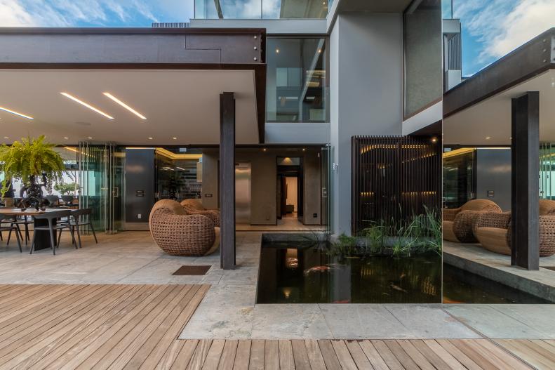 A wood walkway leads into an indoor-outdoor living area of a modern estate. A pair of woven chairs face an in-ground water feature that houses vegetation and provides relaxation. Tile flooring takes over where the wood-paneled walkway ends, leading into the rest of the home.