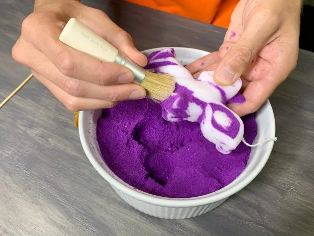 The paraffin wax will slightly shrink when cooling and separate from the sand. Once hardened, carefully remove the candle from the mold. Use a paintbrush with stiff bristles to brush off leftover stubborn sand that is stuck to the candle. Use the wooden skewer to help remove sand from any detailed areas. Carefully break away any wax that over spilled in the mold. TIP: If your candle doesn’t stand, use a sharp knife to carefully cut a straight edge for the base.