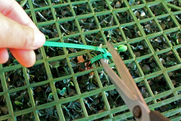 Referencing your design, begin weaving the faux flowers into the boxwood panels using zip ties to secure: Hold the stem in place against the boxwood panel, then weave an end of a zip tie through an opening in the mesh of the panel, over the stem and through the back of the mesh again before securing (Image 1). Cut and discard loose ends of the zip ties (Image 2).