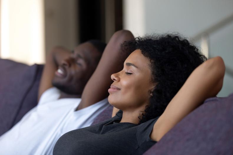 A Black man and woman relax with their eyes closed on a couch.