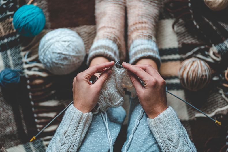 A woman's hands are shown knitting with light-colored yarn.