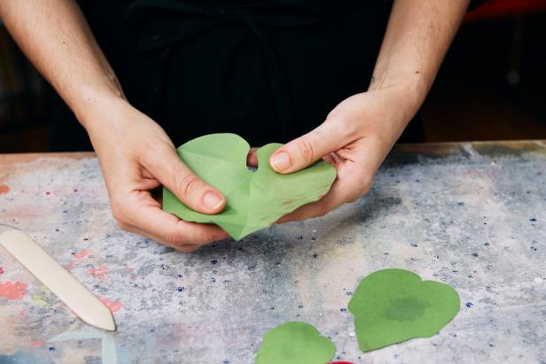 Fold the leaves to mimic vines. Use a bone folder. Fold in half and press it down with a bone folder. Then, create a 45 degree fold and press with a bone folder. Repeat two more times. Open the leaf up and fold the tops inward so the leaves appear like a little curved boat.