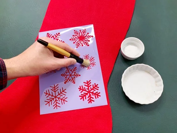 Stencil snowflakes on the top piece of felt. Place the stencil in the desired spots and apply white fabric paint on the brush and dab it off on a separate dish. This will ensure that there isn’t too much paint on the brush. Start towards the outside of the stencil and work into the center in an up and down dabbing motion. Don’t worry about imperfections; this will add a vintage vibe. Continue this process around the stocking until you’re happy with the look. Let the paint fully dry before moving on to the next step. Tip: Use a cutting machine to cut out a personalized stencil for a truly custom look.