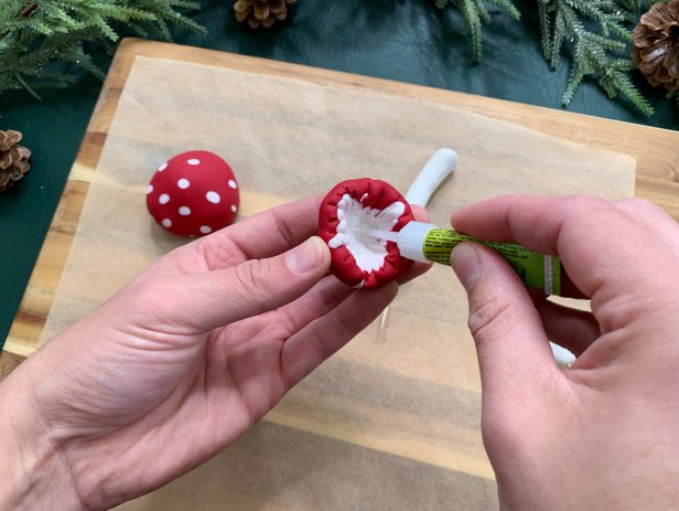 Once baked and cooled, add a little super glue to the bottom of the mushroom cap and press the stem on it. Hold it in place until it fully adheres. Place a toothpick into the bottom of the stem in the pre-made hole. If it does not fit tightly, add a little super glue to the tip of the toothpick before sticking it in. Add a glossy top coat to the mushrooms to really make them pop. Sprinkle on glitter to the tops and sides to give it a frosted winter look.