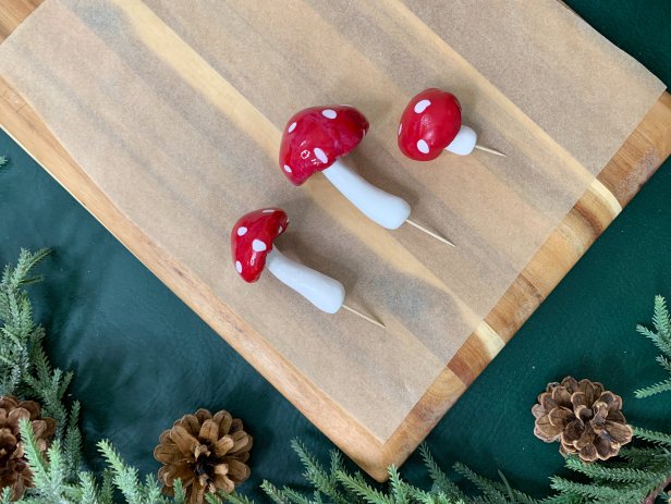 Once baked and cooled, add a little super glue to the bottom of the mushroom cap and press the stem on it. Hold it in place until it fully adheres. Place a toothpick into the bottom of the stem in the pre-made hole. If it does not fit tightly, add a little super glue to the tip of the toothpick before sticking it in. Add a glossy top coat to the mushrooms to really make them pop. Sprinkle on glitter to the tops and sides to give it a frosted winter look.