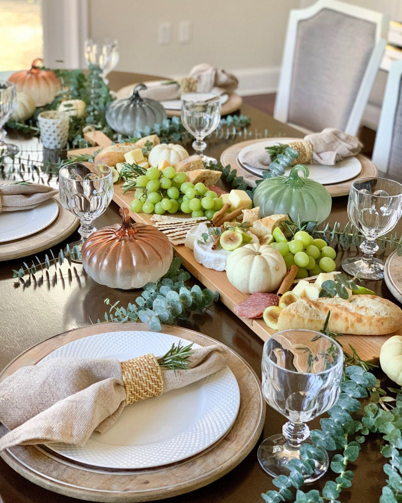 Thanksgiving table with a long wooden charcuterie board centerpiece, glass pumpkins, eucalyptus and linen napkins tied with rosemary