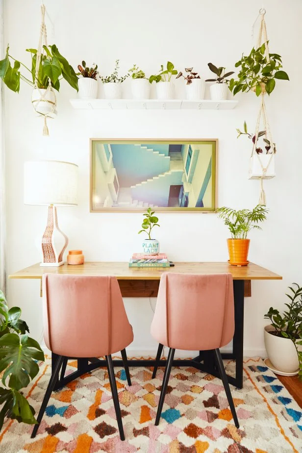 New York muralist and interior decorator Maggie Antalek's lively, retro living room is one that inspires creativity. A floating shelf with houseplants hover above a modern wooden desk with two pink upholstered chairs in the space that doubles as an office. A little pot that says plant lady can be seen on the desk.