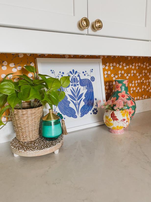 This pantry's countertop is accessorized with colorful new decor as part of its brand new renovation. The renovation introduced color on the walls in the form of a bold new wallpaper in a mustard yellow shade and the new decorations further the colorful aesthetic.