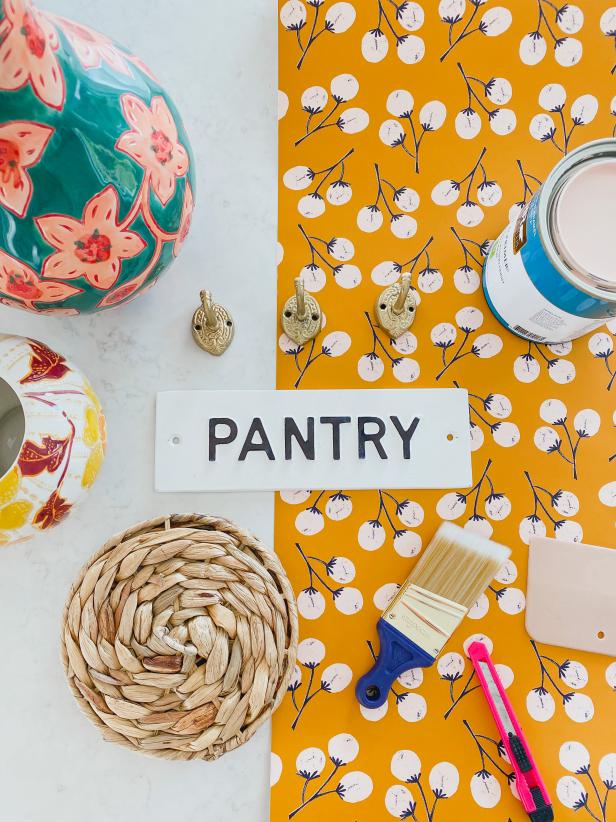 New wallpaper, colorful vases, an enameled sign and other accessories are laying on a table to eventually be installed in a pantry that is receiving a colorful overhaul. The items are grouped to allow a peak into the eventual aesthetic that the pantry will take on.