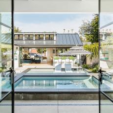 Modern Glass French Doors Open Onto a Concrete Pool Deck That Features Lounge Chairs and a Striped Shade Umbrella