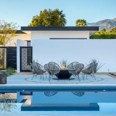An Outdoor Lounge Area Consisting of a Fire Pit and Modern Chairs Sits Beside a Swimming Pool and Between Palm Tree