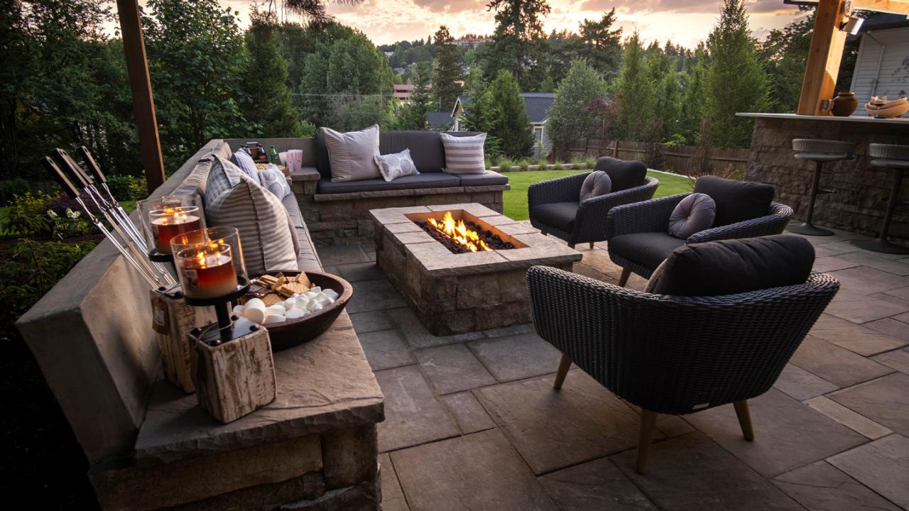 An inviting outdoor patio with a stone fire pit, comfortable seating, and a s'mores setup on a side table, surrounded by lush greenery at twilight.