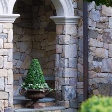 An Ornate Planter, Sitting Beneath a Flagstone Archway, Houses an Evergreen Shrub and a Variety of Flowers