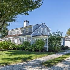 A Cape Cod-Style Home Is Surrounded By Lush Landscaping and Features a Gravel Driveway That Leads to a Garage That Sits at the Back of the Property