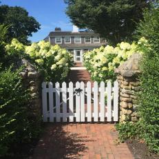 A Brick Walkway Is Surrounded By Lush Landscaping and Leads to the Backyard Though a White Picket Gate