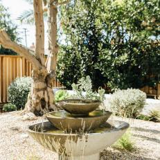 A Stacked Fountain Sits in the Middle of a Gravel-Filled Garden That's Spotted With Lush Vegetation