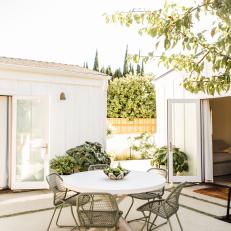 A Modern Outdoor Dining Table and Chairs Sit Between Two White Cottage-Style Buildings