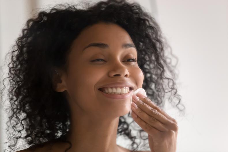 Head shot close up happy african american young woman standing with wide smile, using disposable cotton pads, removing make up before sleeping or cleaning face, applying lotion toner on clean skin.