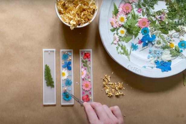 To make a DIY resin bookmark, first trim the flowers to fit the bookmark mold and place them into the molds with tweezers. Arrange the floral design in the molds before adding resin. Then, accent the bookmarks with specks of gold flakes.