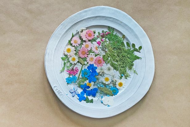 A plate of colorful, dried flowers rests on a work table. To make a DIY resin bookmark, first trim the flowers to fit the bookmark molds with scissors.