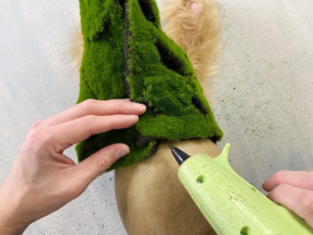 Slide each ear through the cut slits of the moss hat and wrap the two corners around to the back of the egg. Turn the egg over and glue the edges of the hat to the egg. Hot glue the corners and seams together creating a cone. Tip: Keep checking the front of the egg and manipulate the hat as you glue to get the perfect shape.