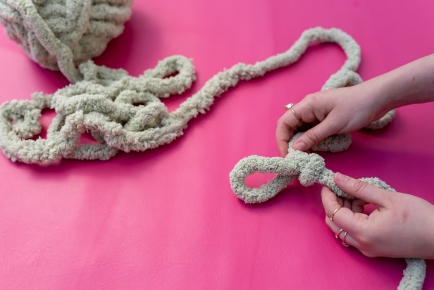 Roll out yarn and leave about 7’’ for the tail and put that to the side. Make the first loop. Fold the yarn over itself to make a loop, then push the extra yarn through the hole and tighten the loop into a knot. Tip: The tightened loop hole should allow about three fingers in width. Next, take the working yarn and put it through the loop to make another one; continue steps to create 15 total loops that make the chain.