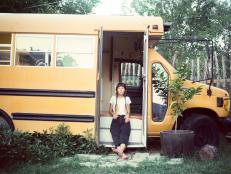 Woman Sits on Steps of Small School Bus in Yard Looking at Camera